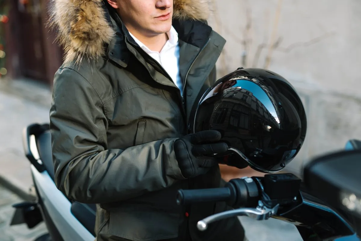 Homme en parka tenant dans ses mains gantées un casque jet noir brillant, debout près de sa moto en ville